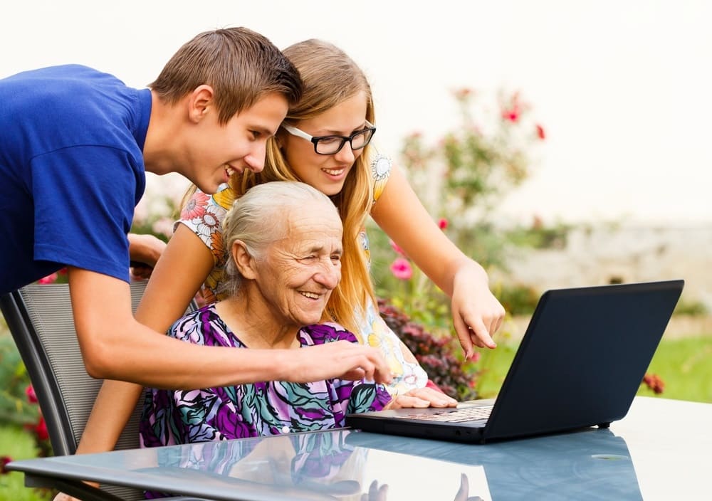 Dois netos, sendo um menino e uma menina, ensinando a avó a mexer no computador. Eles estão do lado externo, em um quintal gramado e todos sorriem. Imagem para ilustrar a matéria sobre avós e netos.