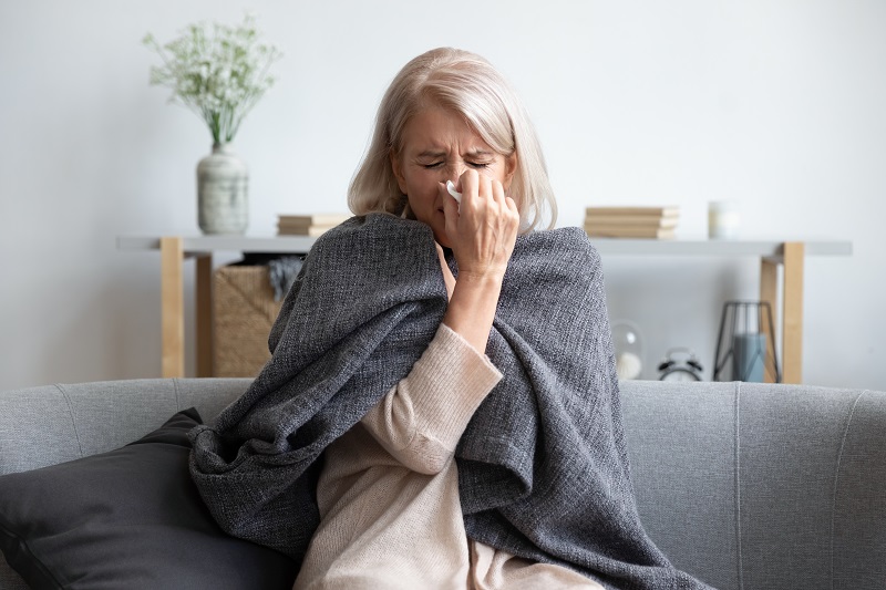 Mulher que aparenta ter mais de 60 anos sentada no sofá, enrolada em uma manta e assoando o nariz, dando a entender que está gripada por causa da baixa imunidade.