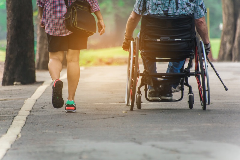 Duas pessoas caminham lado a lado em uma estrada. Uma não aparenta ter deficiência física, enquanto a outra está de cadeira de rodas. Imagem ilustra a notícia sobre isenção do Imposto de Renda para pessoas com deficiência física.