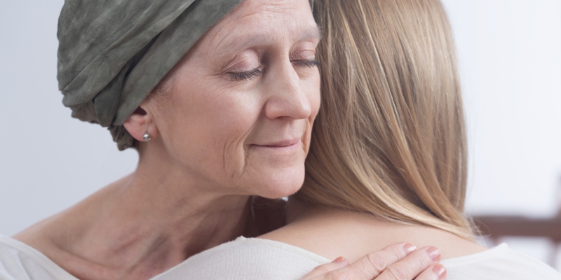 Uma mulher com câncer e lenço na cabeça abraçando uma jovem mulher.