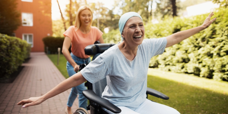 Um jovem mulher levando uma senhora com câncer que está de cadeira de rodas, sorrindo e com braços abertos.