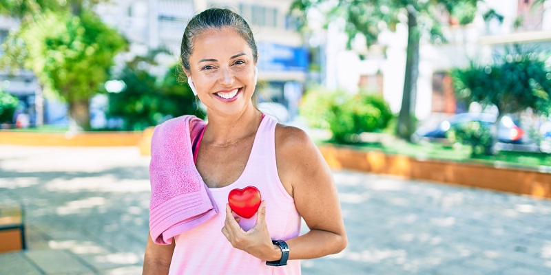 Mulher de 50 a 60 anos se exercita em uma praça, ao ar livre. Ela olha para a câmera e segura um coração em frente ao peito. Foto ilustra o artigo sobre efeitos do envelhecimento no coração.