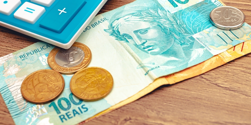 Uma calculadora, uma nota de 100 reais, uma de 20 reais e moedas sobe uma meda. Imagem para ilustrar a matéria sobre o 14º salário do INSS