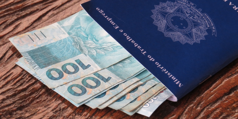 Notas de 100 reais dentro de uma carteira de trabalho. Imagem ilustrativa para a matéria sobre  aposentadoria por contribuição única.