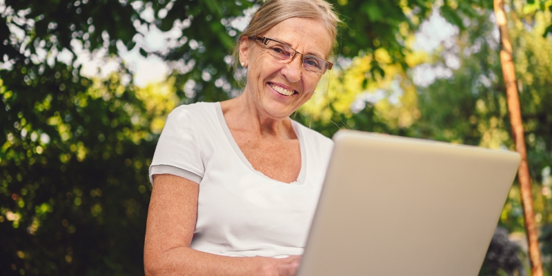 Uma mulher com cerca de 55 anos em um jardim, sorrindo e segurando um computador. Imagem para ilustrar a matéria sobre empreendedorismo feminino.