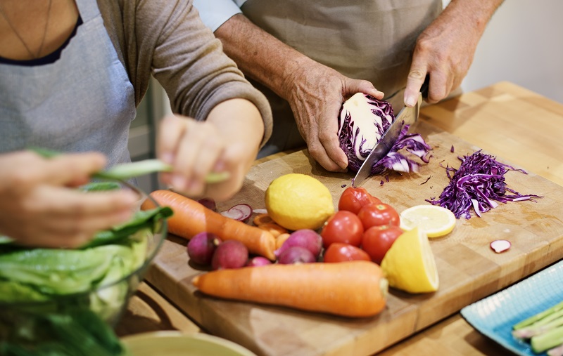 Duas pessoas dividem uma bancada em uma cozinha para cortar verduras, mostrando que alimentação pode ser uma forma para ganhar dinheiro extra.
