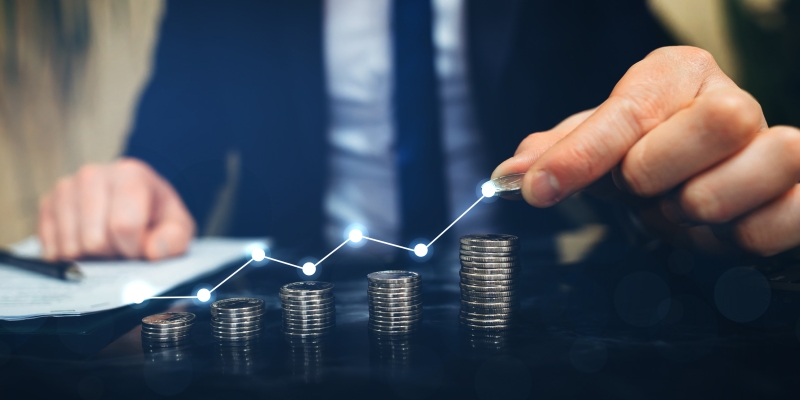 Zoom de moedas sobe uma mesa, com imagem de um empresário empilhando moedas com um gráfico de lucro, mostrando o crescimento de sucesso do negócio. Imagem para ilustrar a matéria sobre poupança.