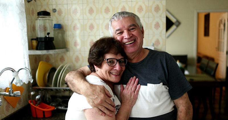 Casal de pessoas 50+ se abraça enquanto olha para a câmera. Eles estão em pé na cozinha de casa. A imagem ilustra o artigo sobre ser velho e não ser doente.