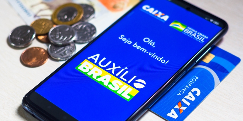 Um celular sobre um cartão da Caixa Econômica Federal e ao lado de moedas. Na tela, o aplicativo do Auxílio Brasil, representando a matéria sobre o pagamento do Auxílio Brasil.