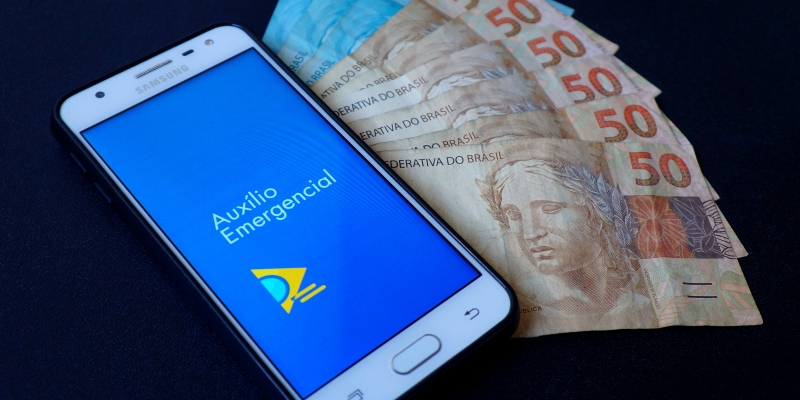 Um celular com a tela do Auxílio Emergencial, sobre notas de 50 e 100 reais. Imagem para ilustrar a matéria sobre pagamento irregular do Auxílio Emergencial.