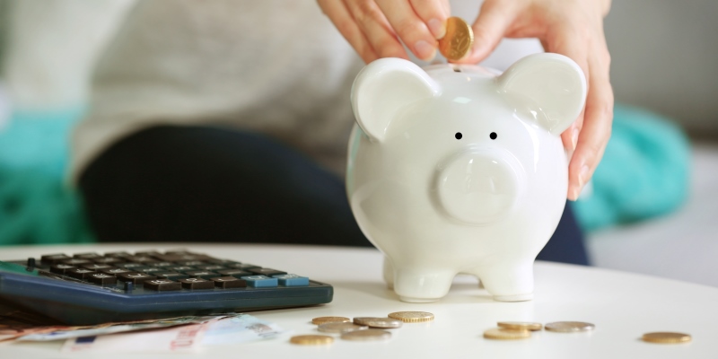 Um mão feminina colocando moedas em um porquinho. Imagem para ilustrar a matéria sobre como economizar dinheiro no dia a dia.