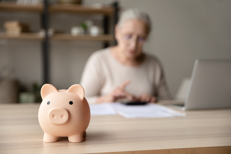 Uma mulher em segundo plano faz contas financeiras. Em primeiro plano, um cofre em formato de porquinho. A imagem ilustra o artigo sobre proteção previdenciária.