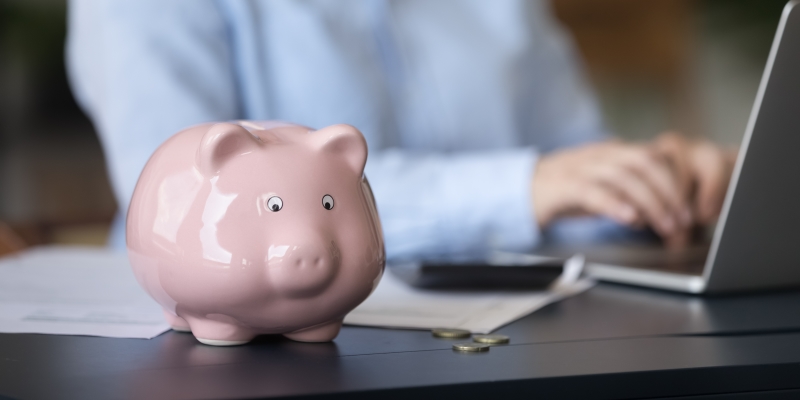 Uma pessoa mexendo no computador. Em cima da mesa, moedas e cofre de porquinho rosa. Imagem para ilustrar a matéria sobre restituição do Imposto de Renda.