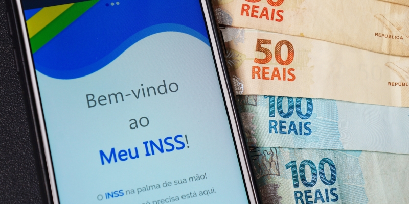 O aplicativo do INSS aberto em um celular. O celular sobre notas de 50 e 100 reais. Imagem para ilustrar a matéria sobre o pagamento da primeira parcela do 13º de aposentados.