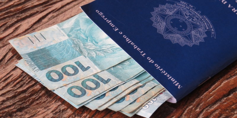 Uma carteira de trabalho com notas de R$ 100 dentro. Imagem para ilustrar a matéria sobre salário dos aposentados do INSS
