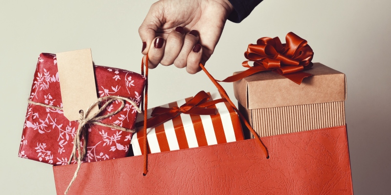 Close da mão feminina segurando um saco de compras vermelho cheio de presentes embrulhados em diferentes papéis. Imagem para ilustrar a matéria sobre décimo terceiro.