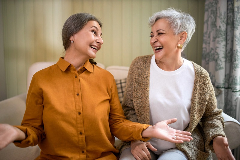 Duas mulheres com mais de 60 anos sentadas em um sofá, lado a lado, conversam e riem.