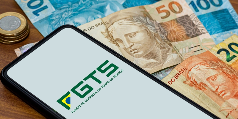 Logotipo do FGTS na tela do smartphone com várias moedas e dinheiros ao redor. Imagem para ilustrar a matéria sobre FGTS extraordinário.