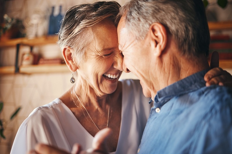 Um casal idoso e caucasiano está abraçado. Com as cabeças encostadas, sorriem um para o outro de olhos fechados.