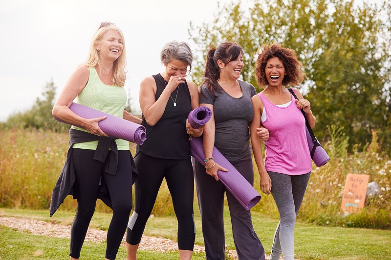 Como viver melhor: 15 hábitos para ter mais qualidade de vida no envelhecimento. Na imagem, quatro mulheres entre 40 e 50 anos caminham lado a lado em um campo. Elas usam roupas de atividade física e carregam tapetes de ioga, enquanto conversam e riem entre si.