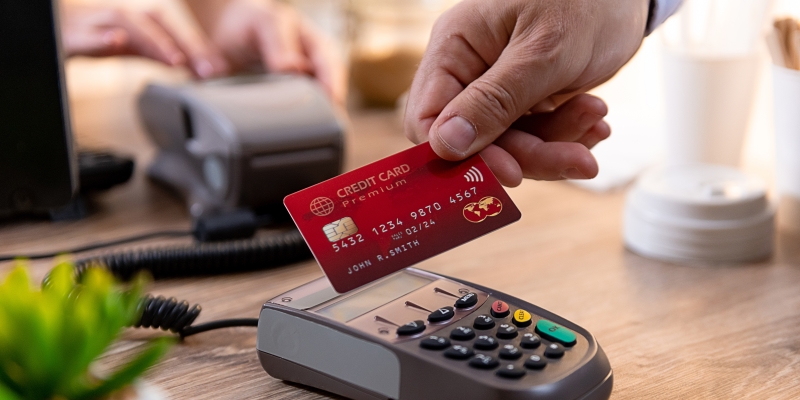 Close de uma mão segurando um cartão de crédito e pagando uma conta ao encostar o cartão em uma maquina de pagamento. Imagem para ilustrar a matéria sobre gastar muito dinheiro.