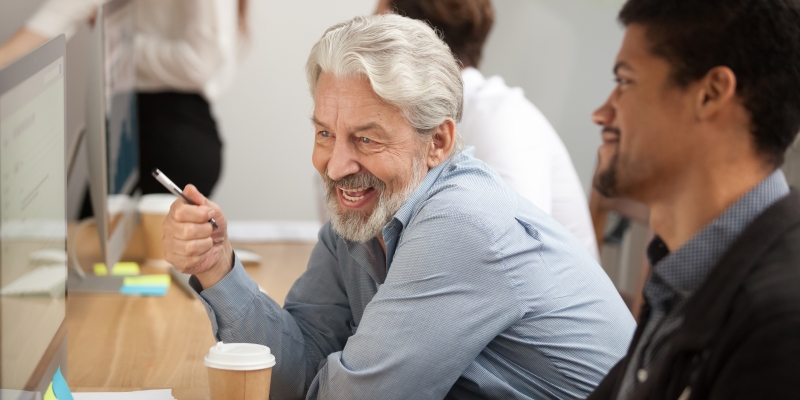 Funcionário sênior sorrindo discutindo e-mail com colega mais novo no local de trabalho. Imagem para ilustrar a matéria sobre envelhecimento ativo.