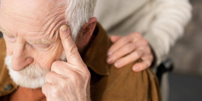 Uma mulher tocando o ombro do marido, doente e com risco de demência, sentado com os olhos fechados e tocando a cabeça.