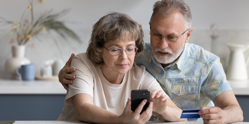 Um casal de senhores olhando o celular, enquanto seguram um cartão de banco. Imagem para ilustrar a matéria sobre banco digital.