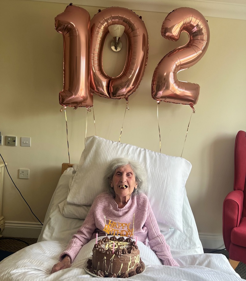 Joyce Jackman, com balões com o número 102 e seu bolo de aniversário, comemorando no lar de idosos com um grande sorriso.