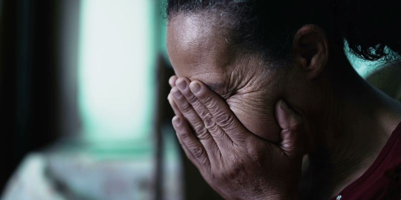 Uma triste mulher idosa cobrindo o rosto com as mãos, sentindo tristeza. Imagem para ilustrar a matéria sobre violência contra a pessoa idosa.