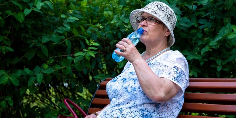Um senhora tomando água, sentada em um banco de praça. Imagem para ilustrar a matéria sobre a importância de tomar água.