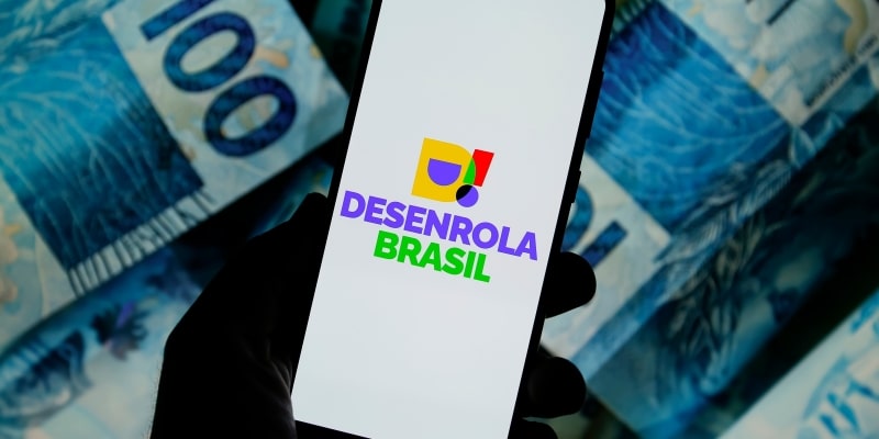 Uma pessoa segurando o celular com a logo do Desenrola Brasil. Ao fundo, notas de R$ 100. Imagem para ilustrar a matéria sobre o golpe do Desenrola Brasil.