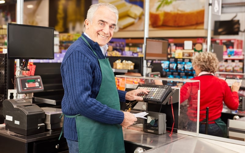 Um senhor de mais de 60 anos trabalhando como caixa de supermercado. Imagem para ilustrar a matéria sobre idade ideal de aposentadoria.