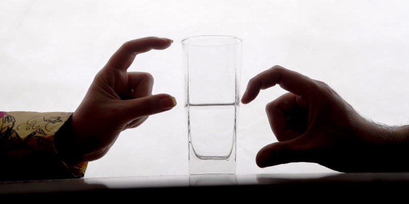 Dois pares estão mostrando metade cheio e metade vazio lado de copo de água com os dedos. Imagem para ilustrar a matéria sobre otimismo.
