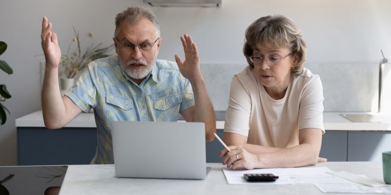 Um casal de cerca de 50 anos, com um computador, uma calculadora e contas a pagar. O marido briga com a mulher. Imagem para ilustrar a matéria sobre abuso financeiro.