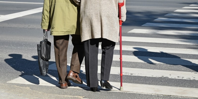 Um casal de idosos atravessando da rua. A mulher anda com uma muleta. Imagem para ilustrar a matéria sobre cidades preparadas para o envelhecimento da população.
