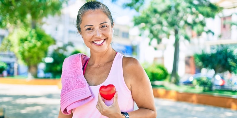 Um mulher de meia idade, após fazer exercício físico, segurando um coração em frente ao peito. Imagem para ilustrar a matéria sobre hábitos hábitos que retardam o envelhecimento.