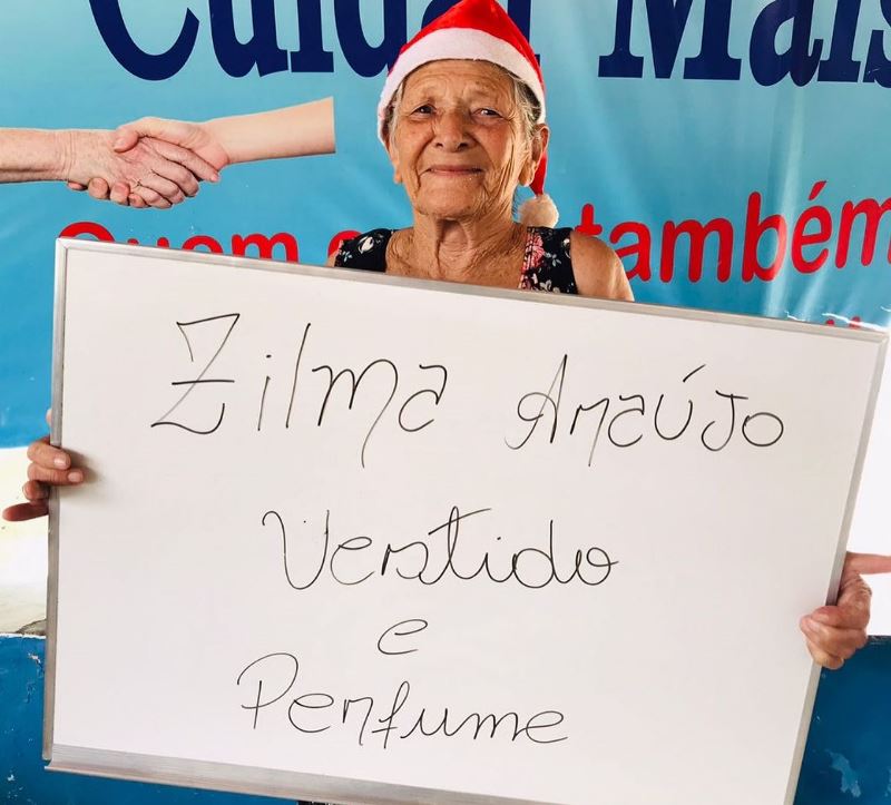 Uma senhora, com uma plaquinha, pedindo presentes simples de Natal, como vestido e perfume. O nome da senhora é Zilma Araújo.