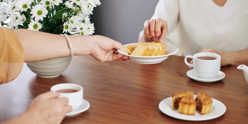 Mulher idosa oferecendo prato com biscoitos para seu convidado na mesa da cozinha enquanto eles estão tomando chá. Imagem para ilustrar a matéria sobre atitudes na alimentação.