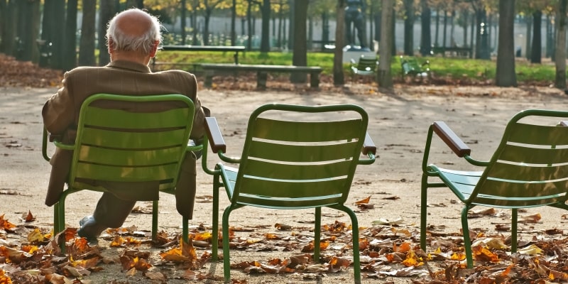 Homem idoso sentado sozinho no banco do parque debaixo de uma árvore e ao lado de duas cadeiras vazias. Imagem para ilustrar a matéria sobre economia da solidão.