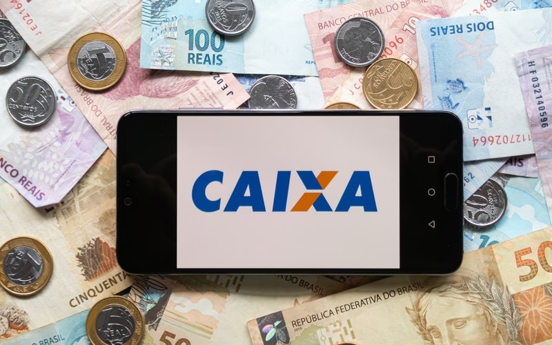 Imagem de um celular com o logo da Caixa na tela. O celular está sobre notas de dinheiro e moedas. A imagem representa a matéria sobre a Mega da Virada.