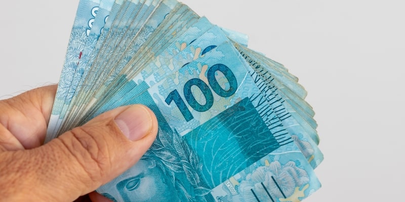 Um mão masculina segurando notas de 100 reais. Imagem para ilustrar a matéria sobre pagamento extra do INSS.