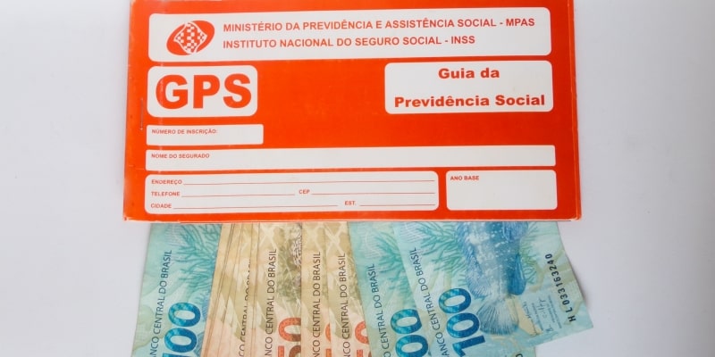 Uma Guia da Previdência Social. Abaixo dela estão notas de 100 reais e notas de 50 reais. Imagem para ilustrar a matéria sobre contribuição de autônomos.