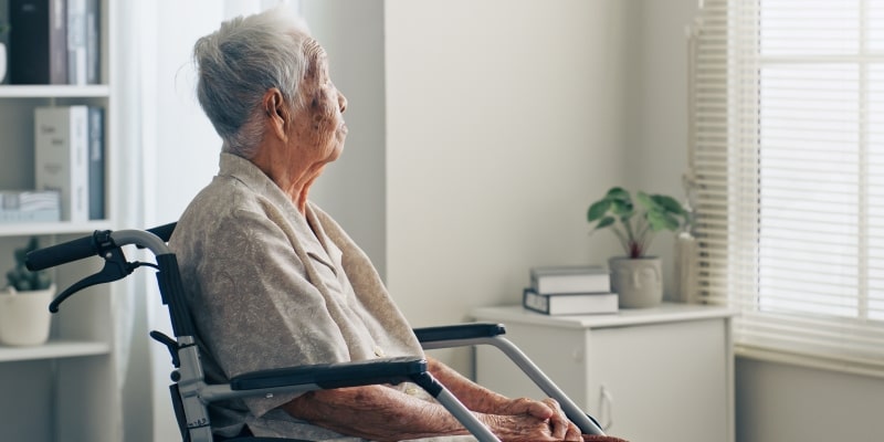 Mulher asiática idosa e solitária, sentada em uma cadeira de rodas, olhando pela janela. Imagem para ilustrar a matéria sobre custos com demência.