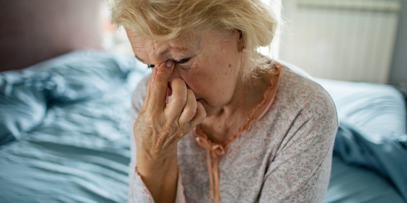 Uma mulher idosa, sentada na cama, com as mãos no rosto e uma expressão de tristeza. Imagem para ilustrar a matéria sobre efeitos da solidão.