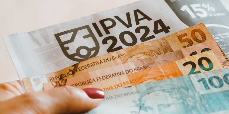 Mãos femininas segurando uma conta do IPVA 2024 e notas de 100, 20 e 10 reais. Imagem para ilustrar a matéria sobre pagar as contas de começo do ano.