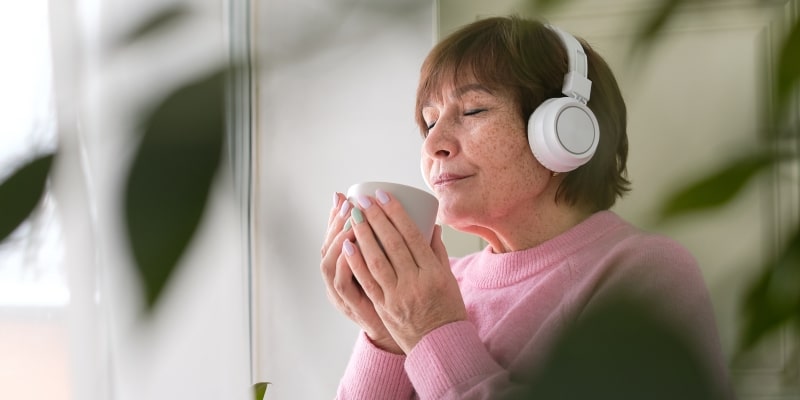Uma senhora idosa perdida em pensamentos junto a uma janela. Ela está com fones de ouvido e, em suas mãos, uma grande caneca de chá. Imagem para ilustrar a matéria sobre solidão ou solitude.
