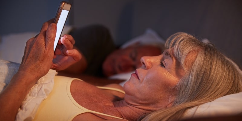 Um mulher deitada na cama e mexendo no celular. Ao lado, está o seu marido, dormindo. Imagem para ilustrar a matéria sobre uso excessivo de telas.