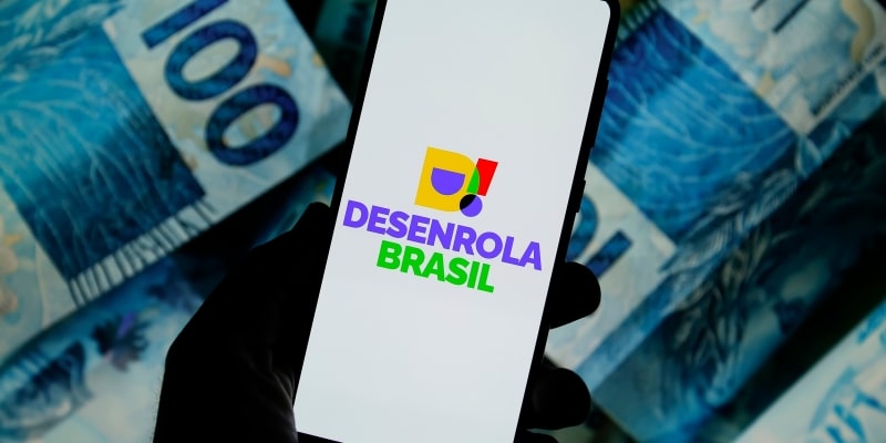 Uma mão segurando o celular com a logo do Desenrola Brasil. Ao fundo, notas de 100 reais. Imagem para ilustrar a matéria sobre Desenrola para empresas.
