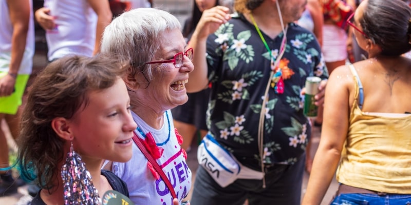 Um senhora curtindo um bloco de carnaval de rua ao lado de sua neta. Imagem para ilustrar a matéria sobre dicas de carnaval para idosos.